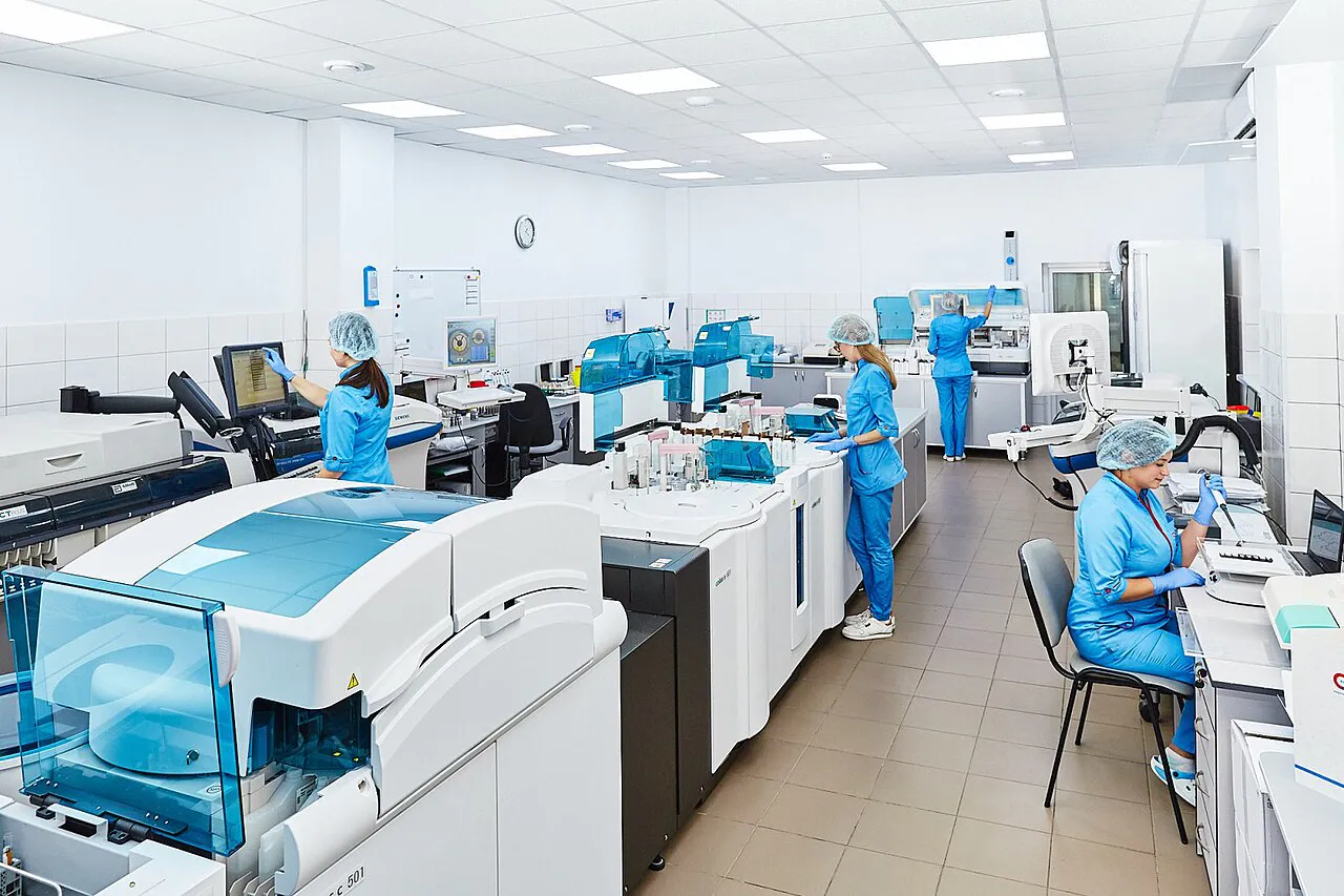 The image shows a group of scientists working in a laboratory. The scientists are wearing lab coats and gloves. They are working on a variety of different tasks