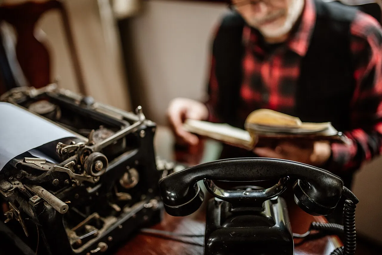 An elderly man reads a book while seated at a desk, surrounded by a vintage typewriter and a classic telephone. The room exudes a warm, inviting atmosphere perf